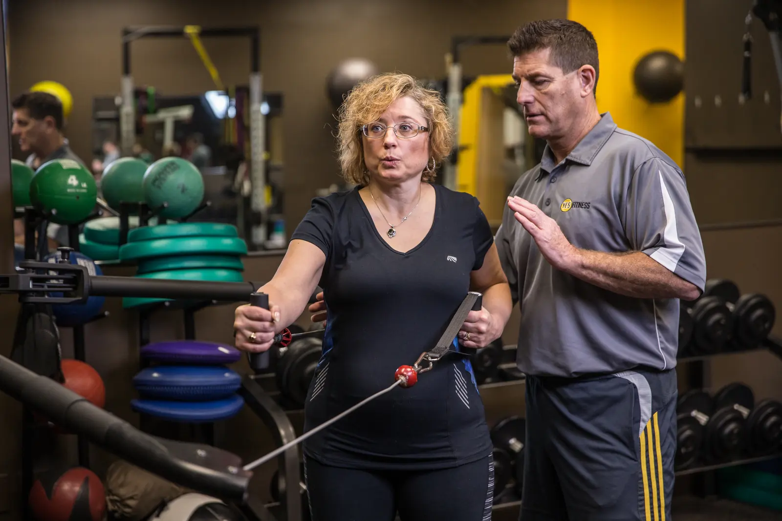 A YES Fitness coach guiding a member through a split-stance cable push pull